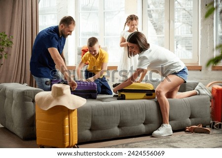 Man, woman, boy and girl sorting garments for travel, nd packaging bags in bright living room. Joyful mood before trip. Family resort. Concept of tourism, holiday, vacation, relaxation. Ad Royalty-Free Stock Photo #2424556069