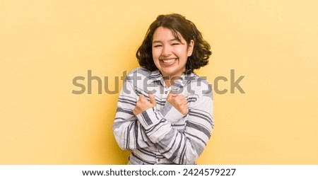 pretty hispanic woman smiling cheerfully and celebrating, with fists clenched and arms crossed, feeling happy and positive