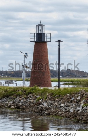 Distant view of small brick light house at water's edge of lake with bench and lamp post under cloudy sky
