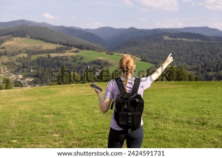 Blond girl with phone pointing finger on mountains background. Young female with backpack hiking in nature. Beautiful woman walking in sport clothes. Freedom and active lifestyle concept, rear view