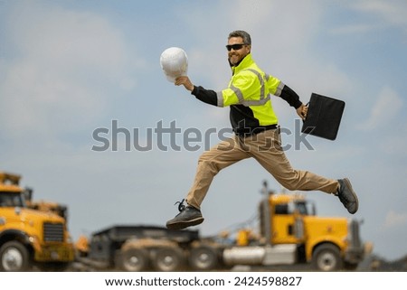 Hispanic builder excited jump and run on site construction. Excited builder construction worker in a safety helmet jumping in front of the trucks. Excited crazy builder man in helmet jump outdoor. Royalty-Free Stock Photo #2424598827