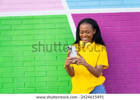 A cheerful African lady in a bright yellow top smiles at her phone, against a colorful geometric background. Royalty-Free Stock Photo #2424615491