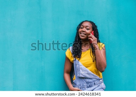 Joyful Latinx lady chatting on a red cell phone, with a bright blue wall backdrop, hand on hip, expressing happiness