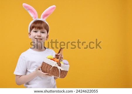 Easter celebration. Cute little boy with bunny ears and wicker basket full of painted eggs on orange background. Space for text