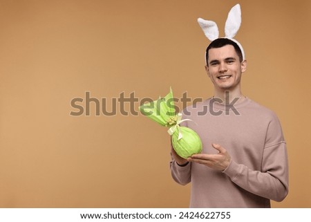 Easter celebration. Handsome young man with bunny ears holding wrapped gift on beige background. Space for text