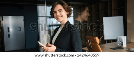 Portrait of saleswoman working in company, standing in office near glass wall, holding mobile phone and smiling, calling client, arranging a business meeting.