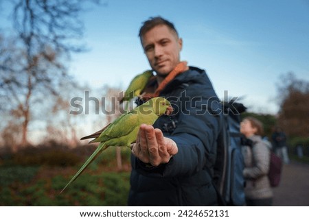 In a London park, a man smiles subtly as a green Roseringed Parakeet perches on his hand, with another on his shoulder, amidst a softlit winter scene with leisurely visitors.