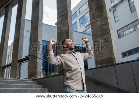 A young happy hispanic man stands outside an office center and courthouse and raises his arms up, rejoicing in success. Royalty-Free Stock Photo #2424655619