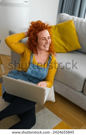 Portrait photo of young smiling woman touching her curly red hair with her hand, while sitting on the carpet and holding laptop in her lap.