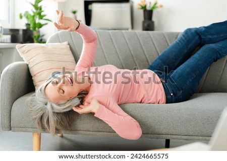 Attractive happy senior woman listening music in wireless headphones, having fun lying on comfortable sofa at home. Technology, positive lifestyle concept 