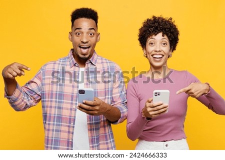 Young couple two friends family man woman of African American ethnicity wear purple casual clothes together hold use point index finger on mobile cell phone isolated on plain yellow orange background