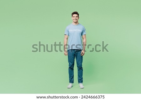 Full body smiling confident fun cool happy young man he wear blue t-shirt title volunteer looking camera isolated on plain green background. Voluntary free work assistance help charity grace concept