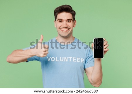 Young man wearing blue t-shirt white title volunteer hold use blank screen mobile cell phone show thumb up isolated on plain green background. Voluntary free work assistance help charity grace concept