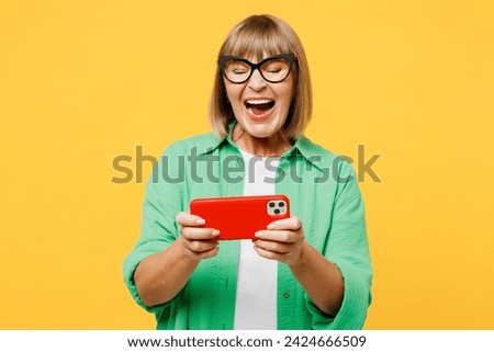 Elderly gambling blonde woman 50s years old wear green shirt glasses casual clothes use play racing app on mobile cell phone gadget smartphone for pc video games isolated on plain yellow background