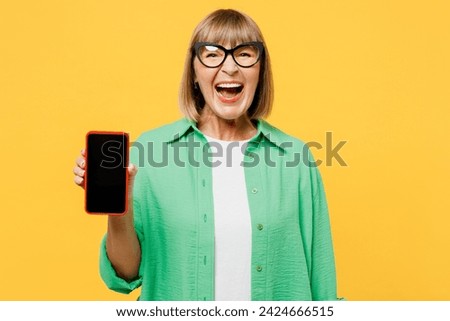 Elderly blonde woman 50s years old wear green shirt glasses casual clothes hold in hand use mobile cell phone with blank screen workspace area isolated on plain yellow background. Lifestyle concept
