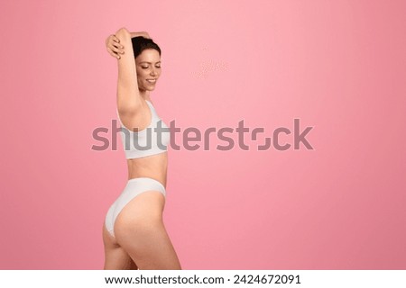 Content and composed, european young woman with closed eyes raises her arms above, showcasing a serene moment in a snug white sports top and bikini on a pink background, studio