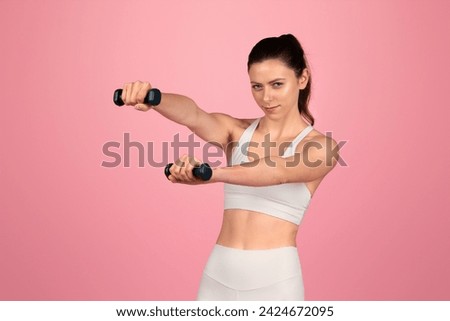 Focused and determined, a woman in a white sports bra and leggings holds out dumbbells with a steady gaze during a workout session, exemplifying strength and fitness on a pink background Royalty-Free Stock Photo #2424672095