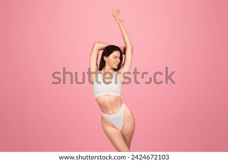 A cheerful european woman with sleek brown hair enjoys a playful stretch in a minimalist white bikini, set against a monochromatic pink backdrop, embodying vitality and joy