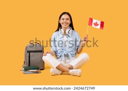 Cheerful young female student holding Canadian flag, with headphones around her neck, sitting with books and backpack on yellow background