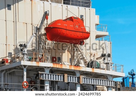 A red lifeboat, securely attached to a large ship by a robust hook, contrasts with the white ship and clear blue sky at a bustling port, promising a safe and adventurous journey. Royalty-Free Stock Photo #2424699785