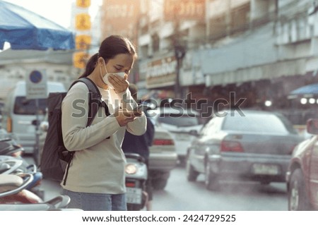 Asian woman wearing PM2.5 dust mask and using phone on the street. wearing mask protect against pollution, anti smog and viruses,  air pollution, and headache suffocating. City air pollution.mist city Royalty-Free Stock Photo #2424729525