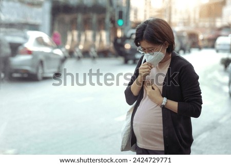 Asian pregnant woman wearing PM2.5 dust mask and coughing on the street. wearing mask protect against pollution, anti smog and viruses,  air pollution, and headache suffocating. City air pollution. Royalty-Free Stock Photo #2424729771