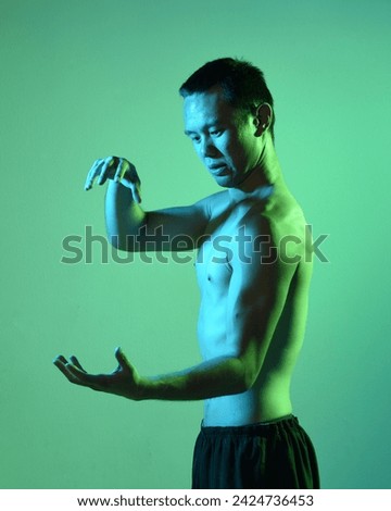 Close up portrait of fit asian male model, shirtless with muscles.  gestural ti chi inspired posing with arms reaching out  Isolated on a moody dark green studio background with silhouette.