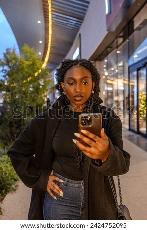 An intriguing scene unfolds as a young Black woman, adorned with elegant earrings and her hair in tight twists, is captured in a moment of connection with her smartphone amidst the urban twilight. The