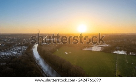 This aerial photograph captures the serene beauty of dawn in a rural setting. The early morning sun breaks the horizon, bathing the landscape in a soft, diffused light. A wind turbine stands in the