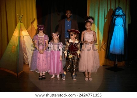 Full length portrait of group of little kids standing on stage in theater with smiling drama teacher Royalty-Free Stock Photo #2424771355