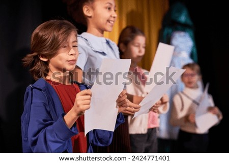 Side view portrait of young boy rehearsing lines standing on stage in school theater in row with children actors Royalty-Free Stock Photo #2424771401