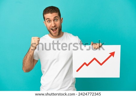 Handsome blonde man over isolated blue background holding a sign with a growing statistics arrow symbol and celebrating a victory