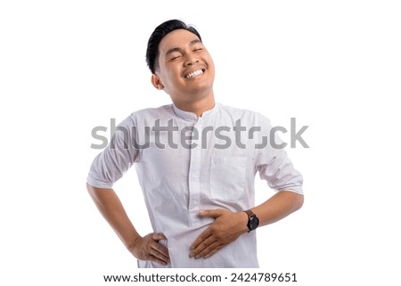 Happy Asian Muslim man touching his stomach feeling full after delicious meal with relieved expression isolated on white background. Ramadan and Eid Fitr celebration concept Royalty-Free Stock Photo #2424789651