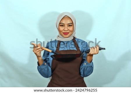 beautiful Asian woman in brown apron and hijab holding spoon and pan while doing homework isolated over white background. muslim housewife concept.