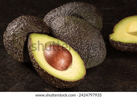Halved avocado next to whole one on black stone table. Dark green skin contrasts with light green, creamy flesh. Nutritious fruit rich in healthy fats, vitamins, and minerals.