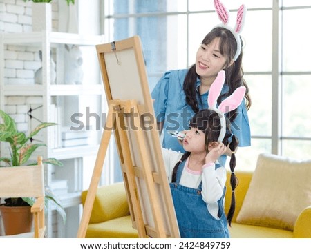 Portrait shot of smiling young asian woman pretty mother helping little cute daughter girl paint picture on canvas having fun together and spending family leisure time in living room at home.
