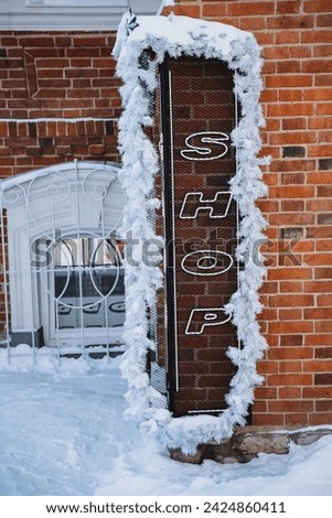 Snow-covered brick wall with a festive shop sign adorned with tinsel. The black metal sign reads shop in white letters, surrounded by snow on the ground. Winter decorations in the city.