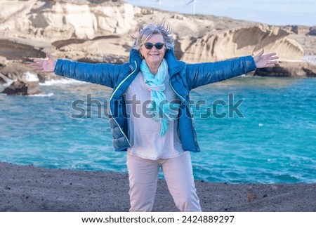 Carefree senior woman enjoying a beautiful windy day at sea, standing on the cliff with open arms. Joyful elderly grandmother appreciating vacation, freedom and retirement