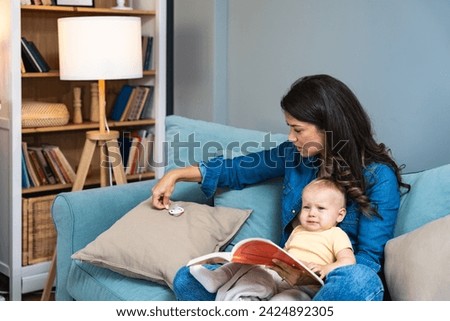 Young mother trying to entertain her newborn baby by reading a fairytale books while sitting on sofa at home. Woman read a book for fun with her little child as a bedtime story.