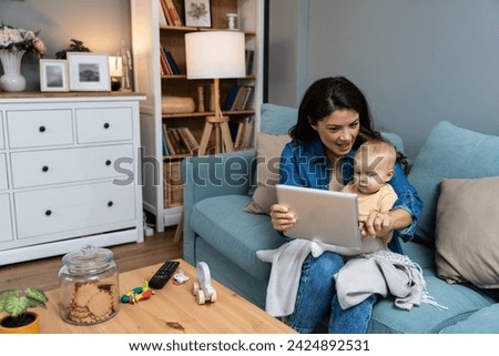 Young mother trying to entertain her newborn baby by playing cartoons and funny videos on digital tablet while sitting on sofa at home. Woman using internet for fun with her little child.