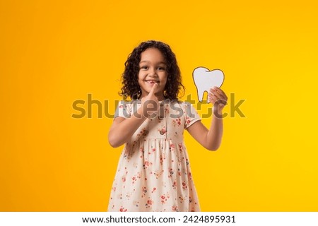 Portrait of child girl holding papercraft tooth and touching her tooth over yellow background. Dental health concept