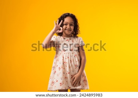 Portrait of smiling child girl showing ok gesture over yellow background. Positive emotions concept