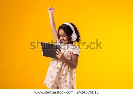 Portrait of smiling kid girl with headphones using tablet and showing winner gesture. Lifestyle, leasure and gadget addiction concept