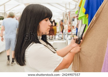 Young Latin Venezuelan woman, at craft fair in Plaza Italia in Buenos Aires, is standing buying clothes and checking prices on mobile application while checking clothes, commerce and tourism concept.
