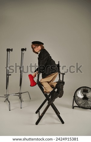 director boy climbing on a director chair and holding red megaphone in hand on grey backdrop Royalty-Free Stock Photo #2424915381