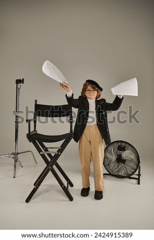 stylish boy in glasses and beret holding screenplay on papers on grey, kid as director of filmmaker Royalty-Free Stock Photo #2424915389