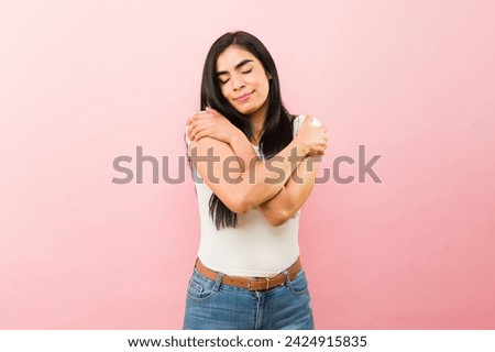 Loving attractive hispanic woman smiling while hugging herself promoting self-love or self-esteem in front of a pink background