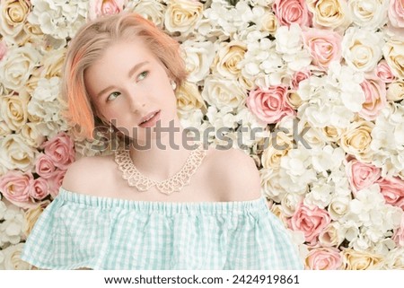 A cute blonde teenage girl with a short haircut in a summer sundress and a pearl necklace poses against a wall of white and pink flowers. Delicate spring-summer look. Beauty, cosmetics. Copy space.