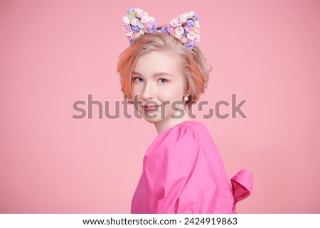 A cute blonde teenage girl with a short haircut poses in a pink dress and a lovely headband with floral kitty ears. Pink background with copy space. Kids and teenage fashion. Spring-summer look.