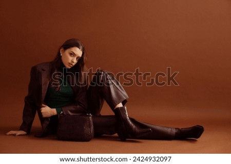 Fashionable confident woman wearing faux leather suit blazer, pants, pointed toe ankle boots, with trendy purple bag, posing on brown background. Full-length studio fashion portrait. Copy, empty space
